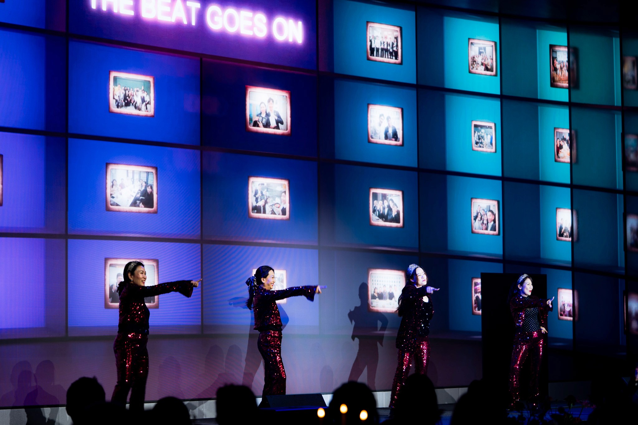Performers in front of a blue tiled LED backdrop displaying photographs and The Beat Goes On.
