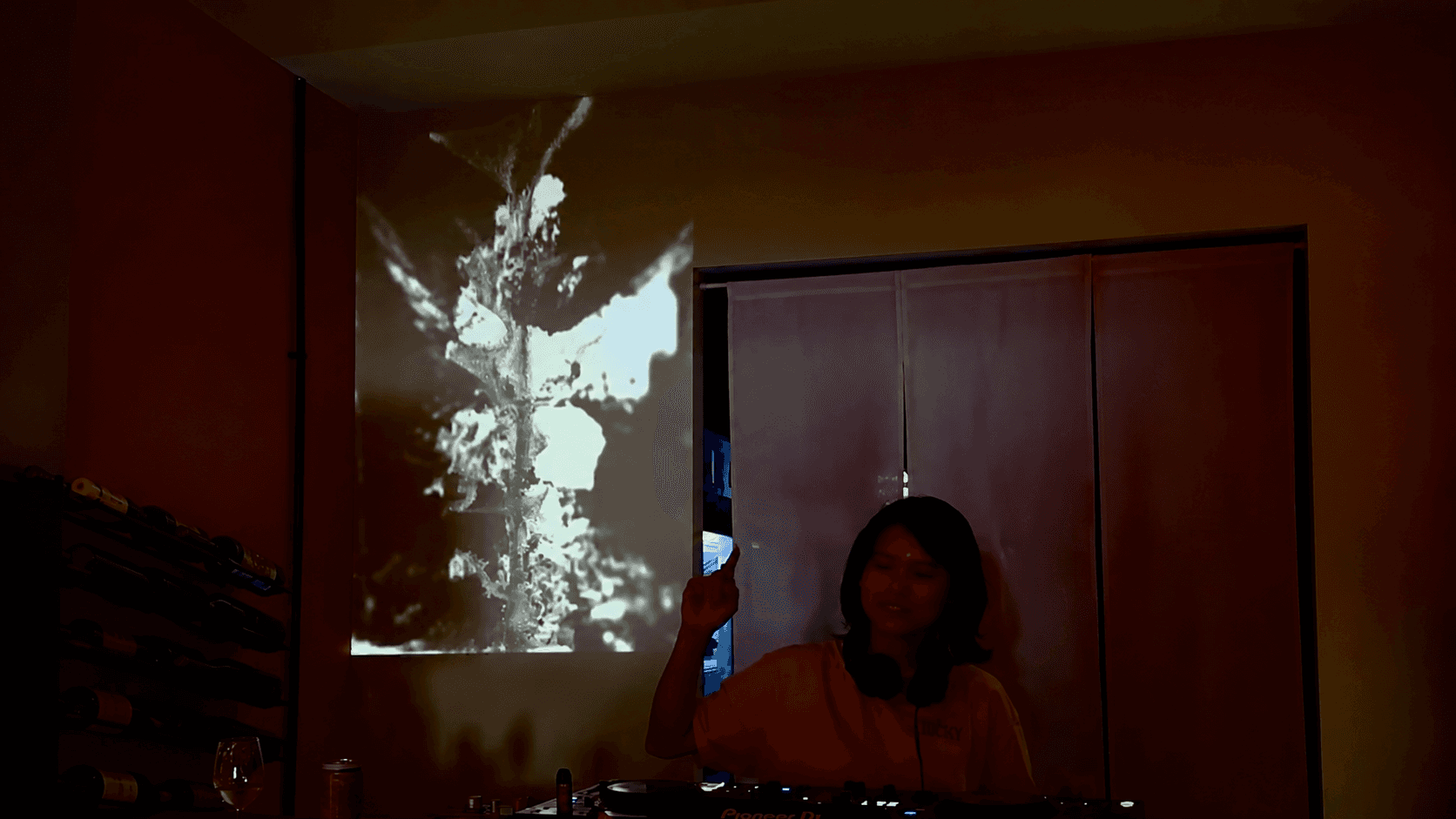 A DJ at a mixing desk in a dim room with abstract white visuals projected on the wall.