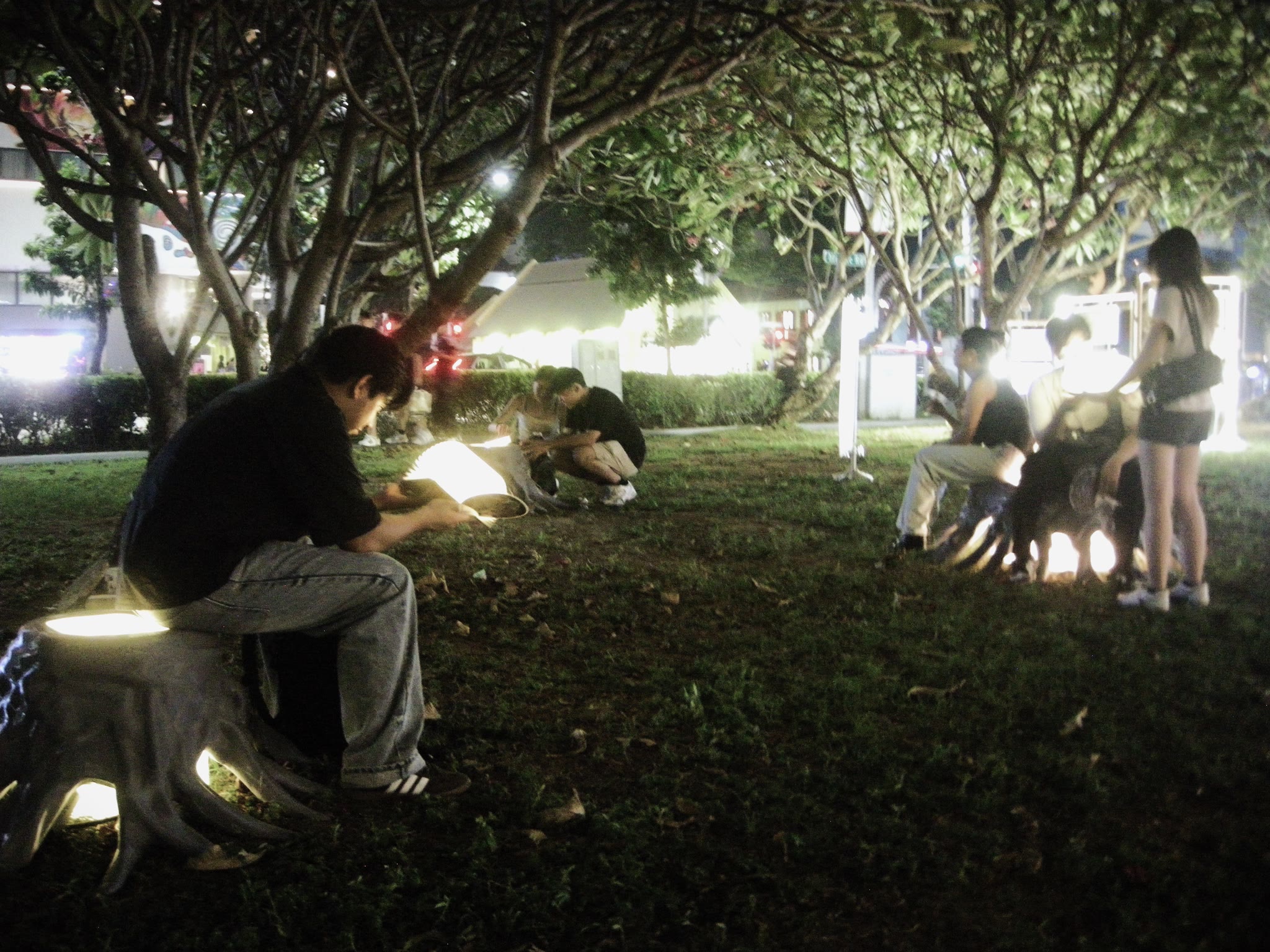 Festival visitors sitting at night on glowing white root-shaped pods under trees, one of them reading an illuminated book.